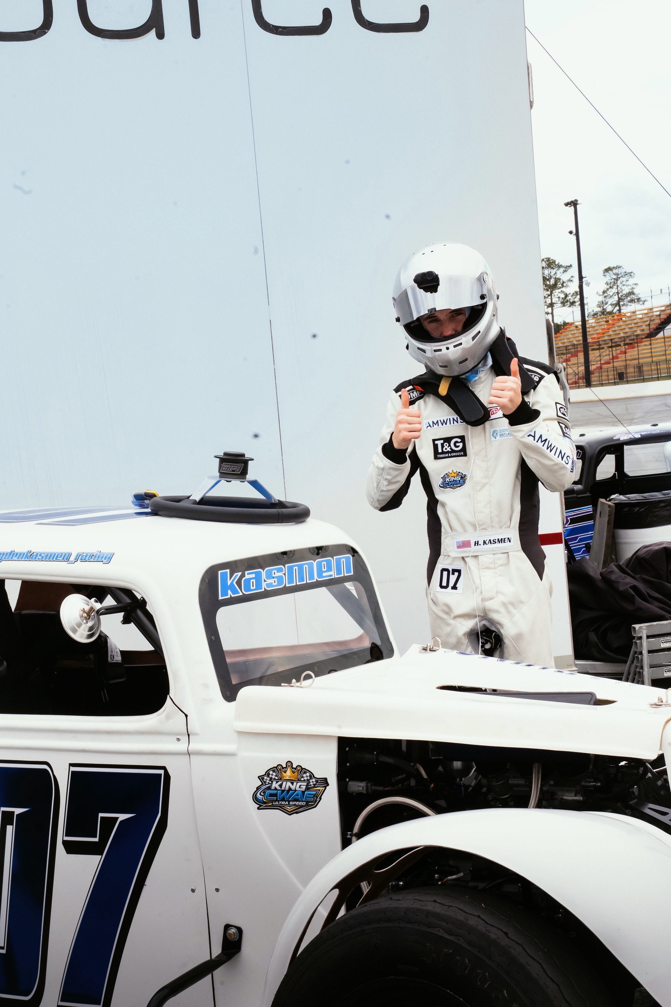 Hayden Kasmen in his racing suit giving a thumbs up next to his No. 07 Legends car